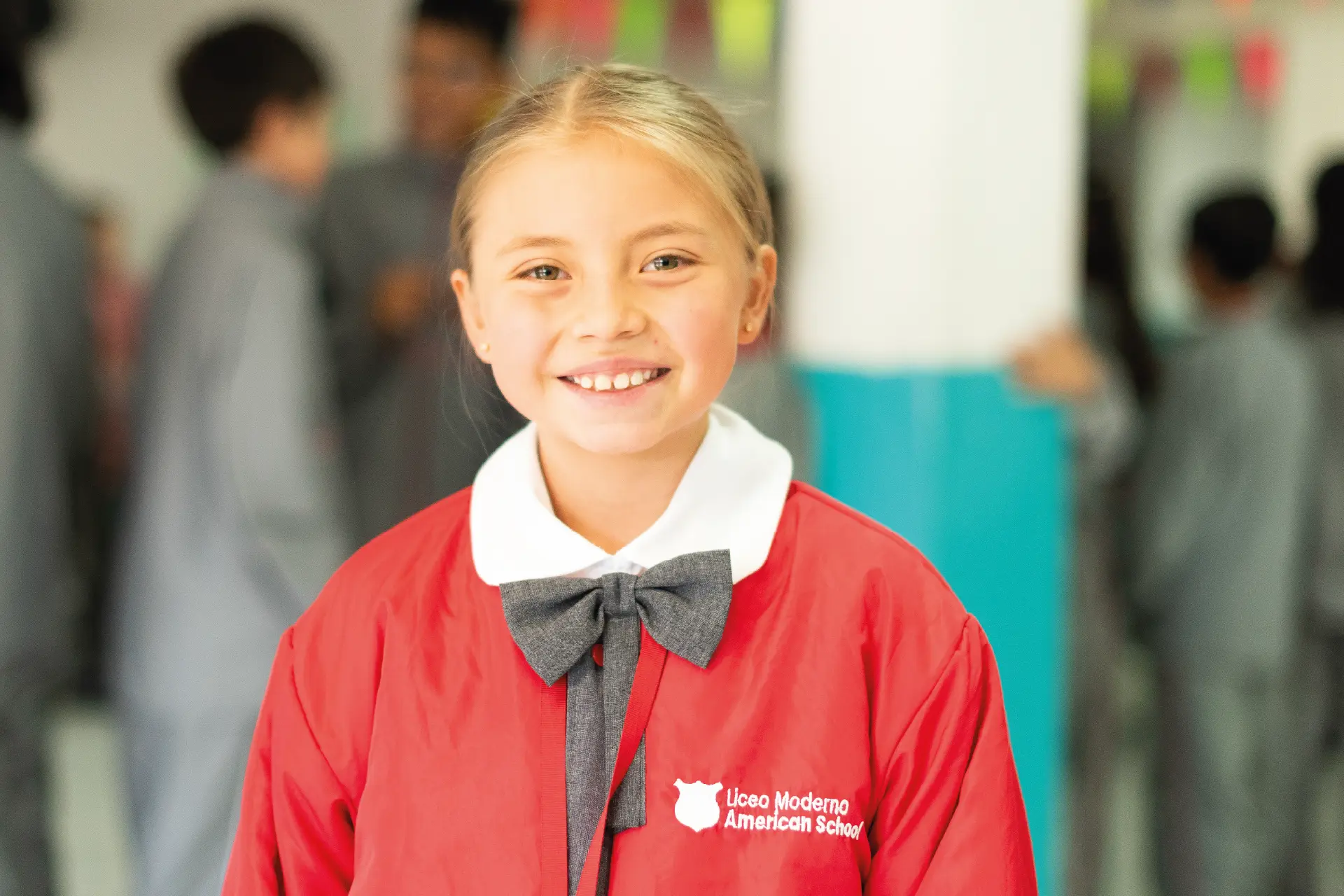 Estudiante del Liceo Moderno American School sonriendo en el pasillo del colegio con uniforme rojo y corbatín gris.